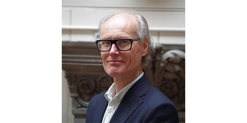 Will Gompertz, wearing a dark blue jacket and a white shirt, stands in the Drawing Office of Sir John Soane's Museum.