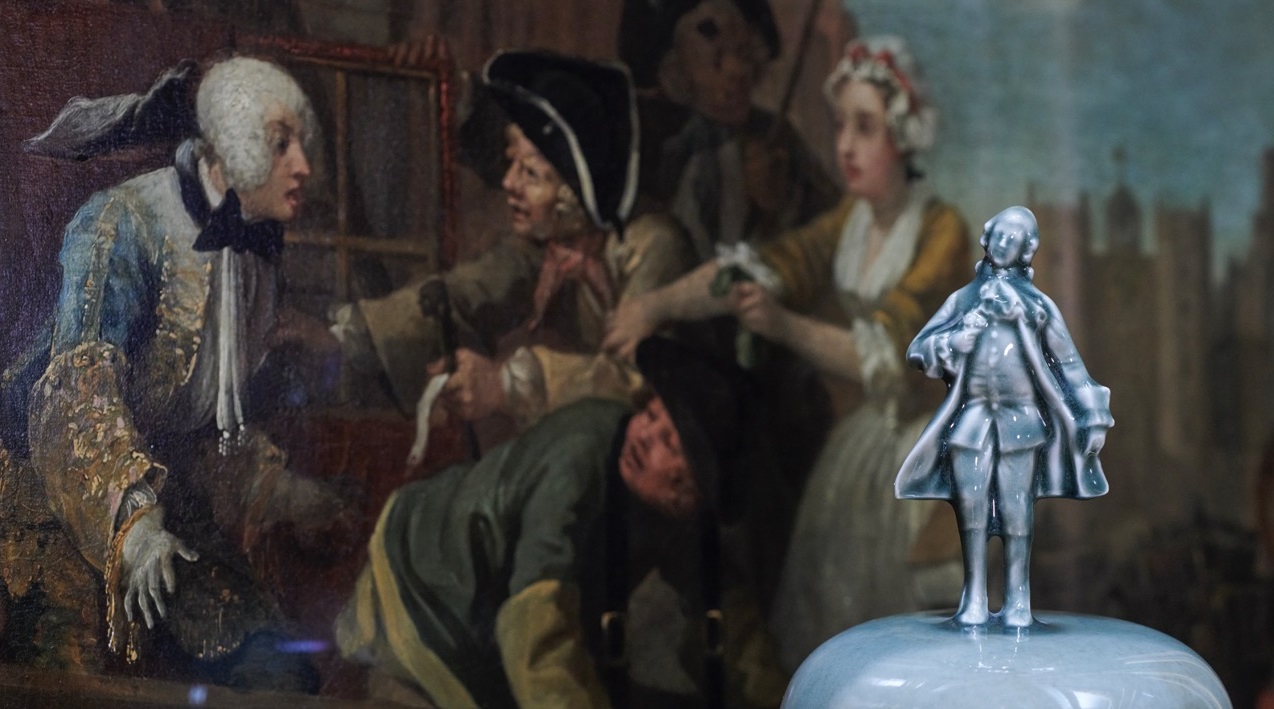A cermaic figure of the Rake standing atop a vase in the foreground, in front of The Arrest, from Hogarth's A Rake's Progress.