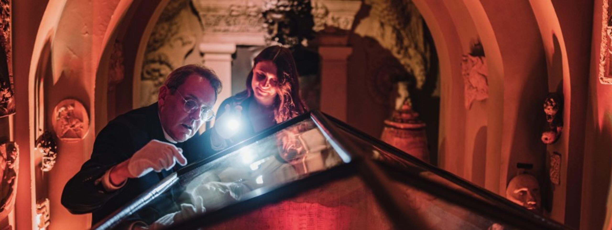 A man shines a torch on a sarcophagus while a young woman looks at it