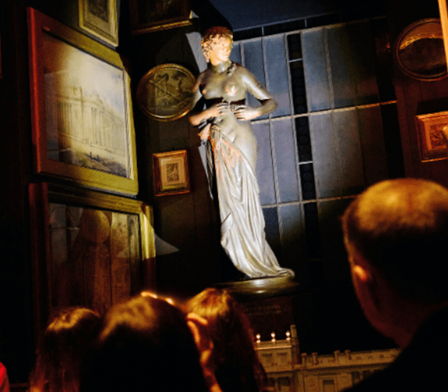 The cast of a Nymph lit by a spotlight in the centre of the Nymph's Recess, surrounded by paintings.