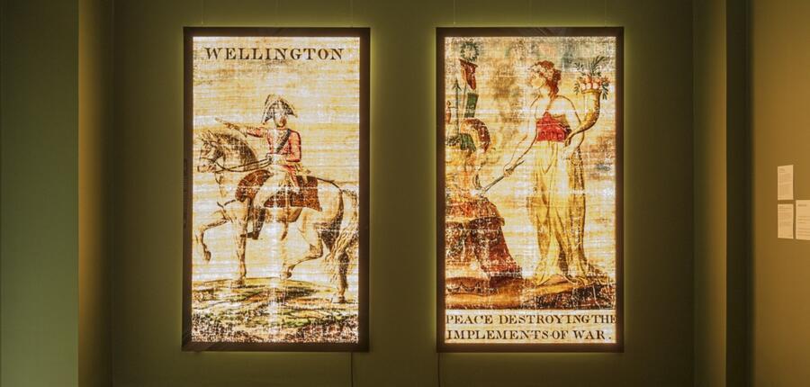 Two illuminated linen transparencies, affixed to light boxes, illustrated with two celebratory scenes from the end of the Napoleonic wars: the Duke of Wellington on horseback, and the figure of Peace destroying weapons.
