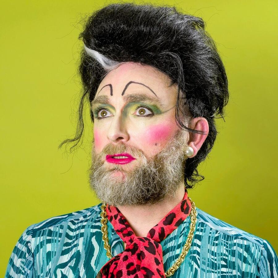Ms. Timberlina; a bearded drag performer wearing a blue patterned shirt and a pink-and-black print shawl.