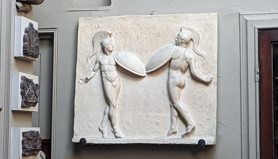 A relief of two gladiatorial male figures, wearing only helmets and carrying swords and shields, as they do battle.