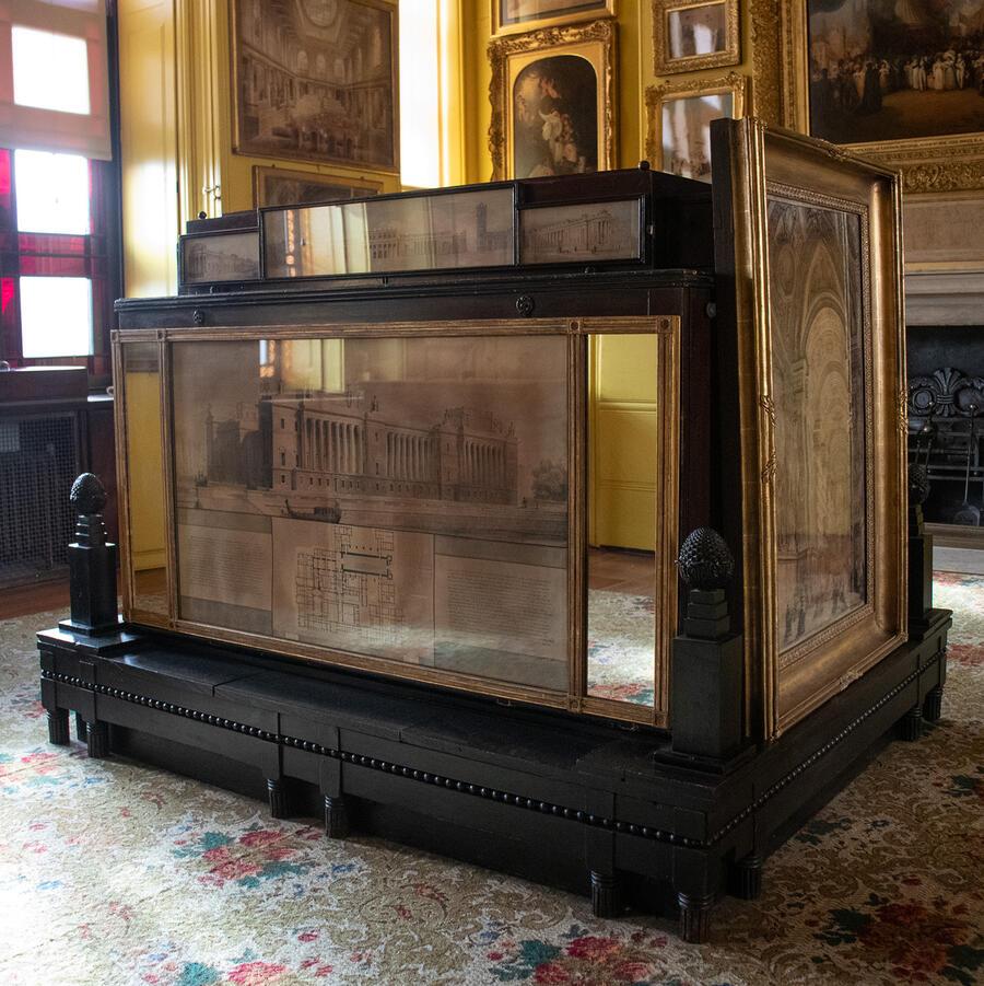 A dark wood cabinet, adorned with framed drawings by George Dance, is positioned at the heart of the Museum's yellow North Drawing Room.