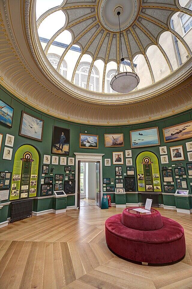 A photo of an open gallery space - a curved wall with a glass dome in the ceiling, and a sofa in the middle. The walls are green and littered with artwork and information