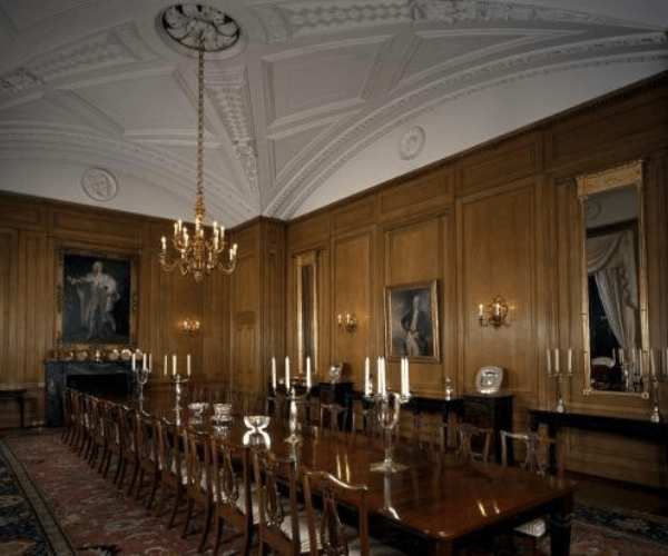 A photo of the State Dining Room at No 10 Downing Street. A large wooden table surrounded by chairs sits in the middle of a yellow room, with ornate chandeliers and candelabra.  