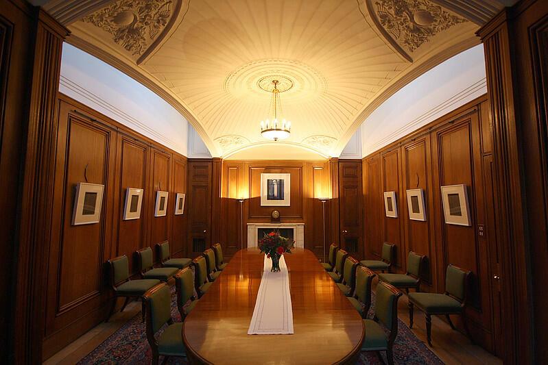 A photo of the State Dining Room at No 11 Downing Street. A large wooden table surrounded by chairs sits in the middle of a wooden panneled room, with a pendentive domed ceiling above, letting in natural light through the sides of the ceiling.