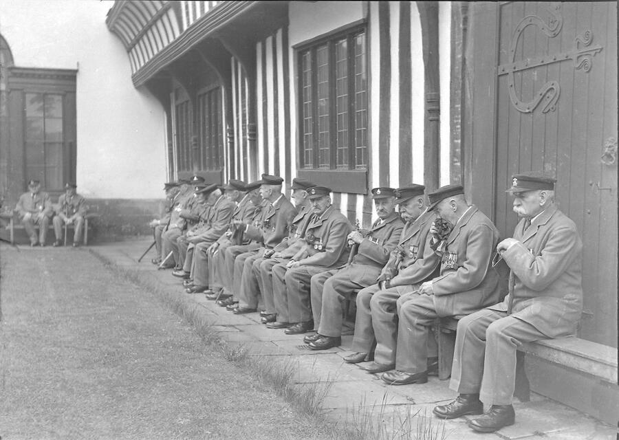 Grey and white photo of elderly pensioners sat on a ledge in a line, infront of a building. The men are all dressed in soldiers uniform, with medals.