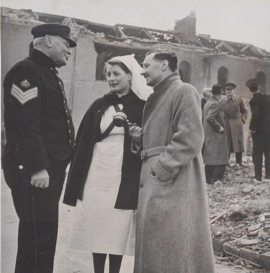 A greay and white photo of a group of people infront of a bombed building. In the foreground one man in a long coat holds a BBC microphone towards a woman dressed in traditional nurses robes.