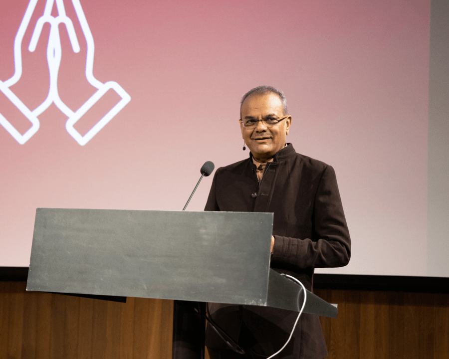 A man stands in front of a podium with a small microphone, and a red screen behind him. He is wearing a suit and smiling.