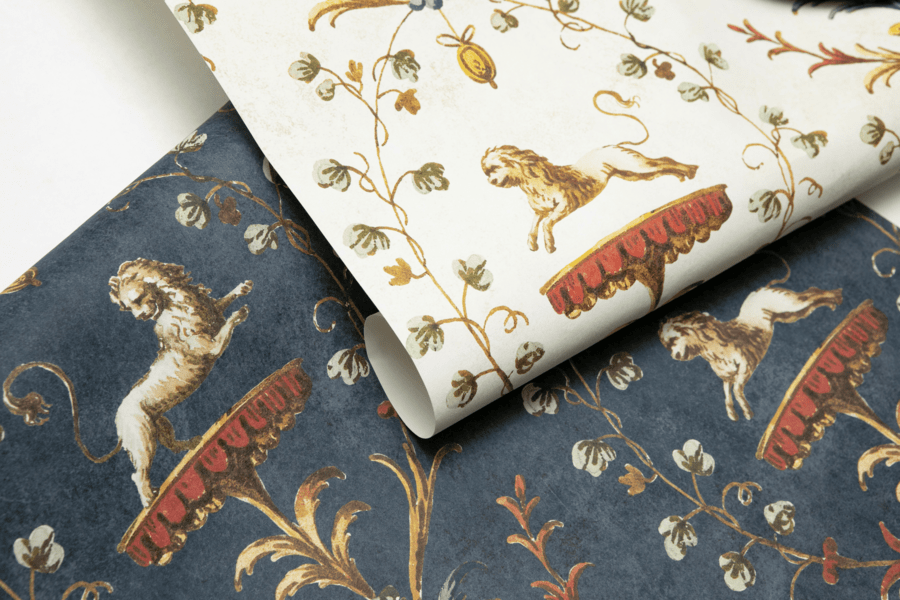 wo sheets of patterned wallpaper shown overlapping. The top sheet has a cream background with a repeating design of golden lions standing on ornate red platforms, surrounded by curling vines with green leaves and small floral details. The bottom sheet has the same design but on a dark blue background, with muted gold and red tones for the lions and platforms. Both patterns have a classical, decorative style.