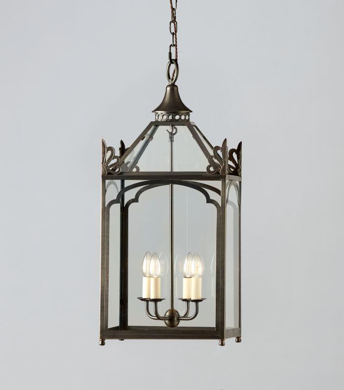 Large bronze hanging lantern with a rectangular glass frame and decorative scroll details at the top corners. Inside, four candle-style light bulbs are mounted on a central stem. The lantern is suspended by a chain against a plain light gray background.