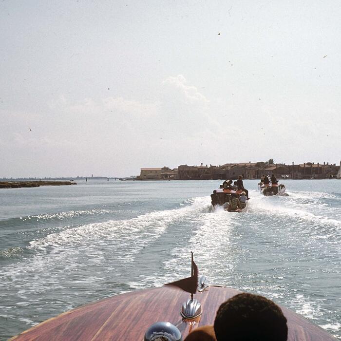 Photo taken from a taxi boat or speedboat in the Venetian lagoon, looking over the bow to more boats ahead and, on the horizon, Venice