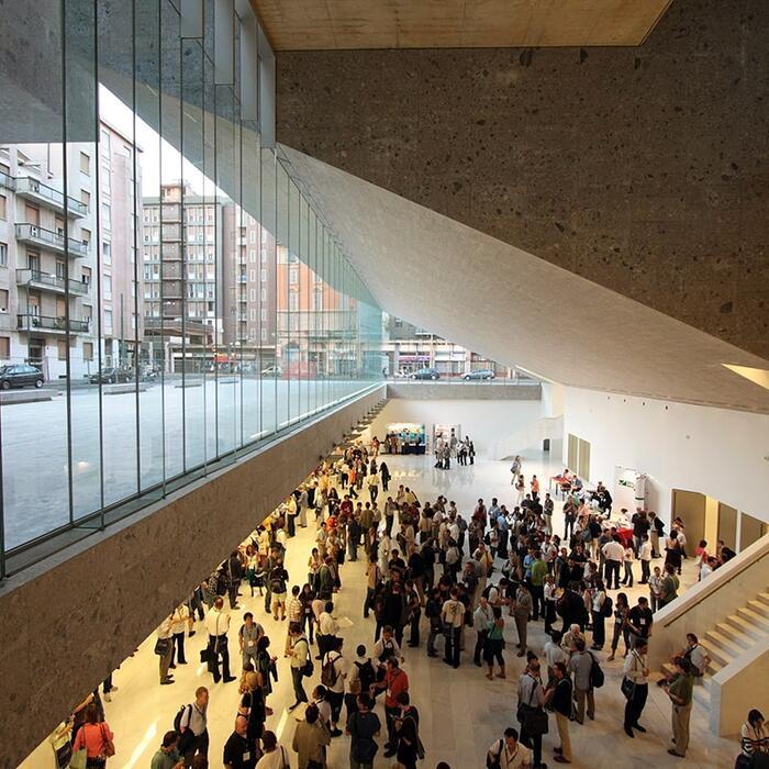 Interior shot of a university building showing lower ground floor hall with students mingling below and windows to the left with a view into the street beyond