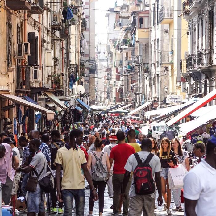 A view down a pedestrianised street in Naples, with crowds moving between shops and cafes, and apartments stretching high above.