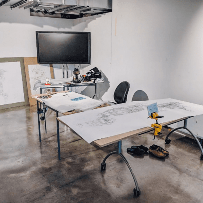 A room with a desk with drawings atop it, a screen and further drawings stacked against a wall