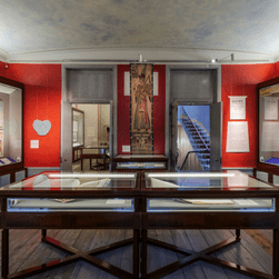 Egypt: Influencing British Design 1775 - 2025 Installation images. Photo shows a red gallery with artwork on the walls, and in glass vitrines.