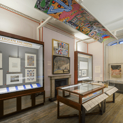 Egypt: Influencing British Design 1775 - 2025 Installation images. Photo shows a white gallery with artwork on the walls, and in glass vitrines.