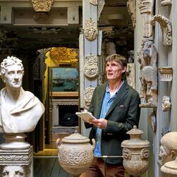 A person stands in an ornate gallery filled with classical plaster casts and sculptures. Visible objects include busts, decorative urns, lion head carvings, and intricate reliefs mounted on the walls. The person is holding an open notebook, and in the background, a framed painting of ships at sea is partially visible through a doorway.