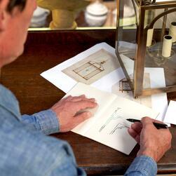 A person is seated at a wooden table, sketching in a notebook with a black pen. The table holds architectural drawings and a brass lantern with candle holders inside. The sketches appear to depict structural or decorative elements.