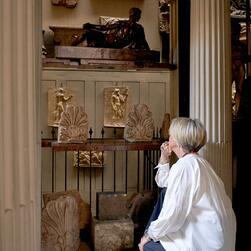 A person is kneeling in front of a display of classical architectural fragments and relief sculptures inside a gallery. The display includes ornate carved panels, decorative motifs, and a reclining figure sculpture positioned on an upper shelf. Two large fluted columns frame the scene.