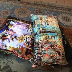 Two decorative cushions placed on a patterned rug. The cushion on the left has an abstract design with vibrant colors including purple, pink, gold, and white, resembling a distorted painting. The cushion on the right features a layered, textured pattern with turquoise, orange, and dark blue tones, incorporating circular and geometric motifs. The rug underneath has muted shades of blue, beige, and rust with floral and medallion designs.