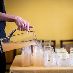A hand pour light coloured juices from a jug against a yellow background