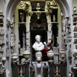 A women stands in the Dome area at Sir John Soane's Museum, surrounded by hundreds of plaster casts. She wears a red apron and holds a duster brush, as she cleans the models.