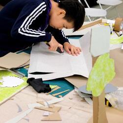 A young boy cuts out a shape from a piece of card, as part of a project to build a cityscape from materials.