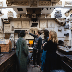 A group of people stand in the drawing office at Sir John Soane's Museum. They are facing towards a man pointing at features on the walls.