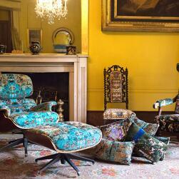 A luxurious interior featuring a yellow-painted room with a white marble fireplace and ornate gold-framed mirror above it. In the foreground, an Eames-style swivel chair with a matching footstool is upholstered in vibrant turquoise fabric with intricate, abstract patterns. Several decorative cushions with similar colorful designs are scattered on the patterned carpet. To the right, an antique high-back chair with dark wood legs and richly patterned upholstery adds a contrasting style. A crystal chandelier h