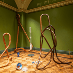 Image of Nika Neelova exhibition install, showing tall wooden sculptures surrounded by lots of colourful glass sculptures in a green room with wooden floors.