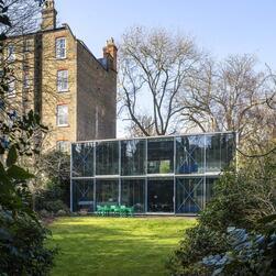 The rear elevation of Hopkins House, as seen from deep in the lush green garden. To the left, a towering Victorian townhouse looms over it.