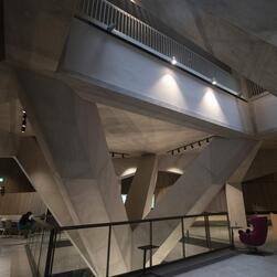 Four huge concrete beams support the above floor, opening a space to the right through which natural light spills from above. Spotlights illuminate the angles of these enormous branches.