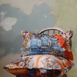 Two decorative cushions stacked on a wooden chair against a textured wall with faded green and blue paint. The cushions feature vibrant, abstract patterns with dominant colors of blue, orange, and red, incorporating circular motifs and layered designs. The chair has a curved backrest with vertical slats, and the overall setting has a rustic, artistic feel.