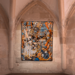 A large, colorful textile artwork displayed on a stone wall within a vaulted architectural space. The artwork features an abstract design with layered patterns in vivid shades of orange, blue, white, and black, incorporating geometric and organic shapes