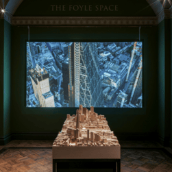 A wooden architectural model showing buildings in London, sits in front of a large screen showing a film.