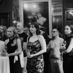 A black and white image of people listening to a speech, drinks in hand