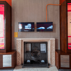 An installation photo of Richard Rogers: Talking Buildings, showing two screens showing a film above a fireplace. 