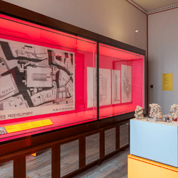 An installation photo of Richard Rogers: Talking Buildings, showing an architectural model on a bright orange plinth in the middle of a room. Behind it is a bright pink wall featuring architectural drawings of the Lloyd's building..
