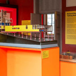 An installation photo of Richard Rogers: Talking Buildings, showing an architectural model on a bright orange plinth in the middle of a room. Behind it is a bright pink wall featuring architectural drawings.