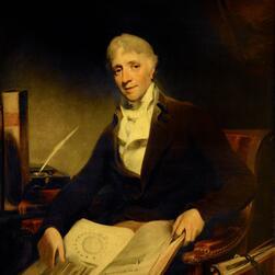 A traditional portrait of a seated figure, Sir John Soane, in formal attire, holding an open book with architectural drawings. The setting includes a wooden chair, a desk with a quill and inkwell, and additional books stacked nearby. The background is dark, emphasizing the subject and the detailed objects in the foreground.