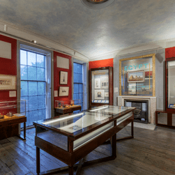 Egypt: Influencing British Design 1775 - 2025 Installation images. Photo shows a red gallery with artwork on the walls, and in glass vitrines.