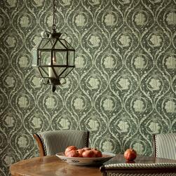 Dining area with a round wooden table, striped upholstered chairs, and a decorative bowl of pomegranates. A geometric glass pendant light hangs above, and the wall behind is covered in dark green damask-patterned wallpaper with a repeating ornate leaf motif