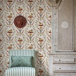 Interior with a striped green and white upholstered chair and a patterned cushion placed against a wall covered in cream wallpaper featuring a repeating design of stylized lions and floral motifs in gold, red, and blue. A large distressed wooden cabinet stands to the right, and a decorative red lion head wall ornament hangs above the chair